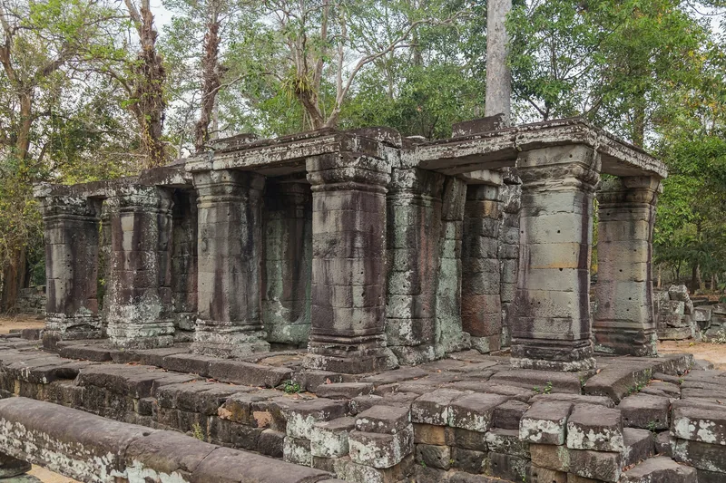 Weathered stone columns and gallery ruins stand among trees at Banteay Kdei temple in Siem Reap, Cambodia.