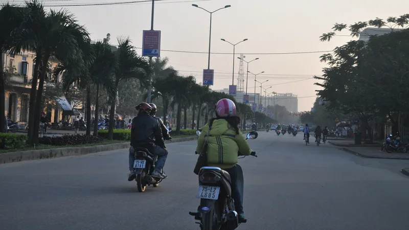 Motorbike riders travel down a palm-lined street in Hanoi under a hazy morning sky.