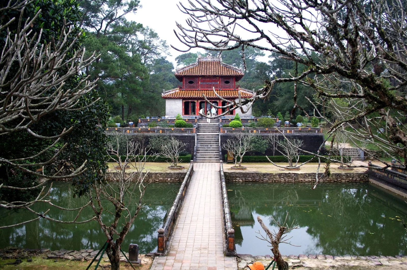 A red pavilion at Minh Mang royal tomb in Hue reflects across a still pond, framed by bare tree branches.