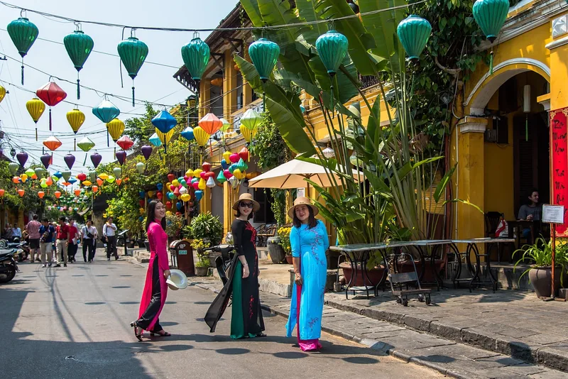 Women in colorful ao dai pose under strings of silk lanterns on a yellow-walled street in Hoi An.